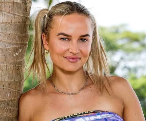 A woman with blonde hair in pigtails smiles, standing by a palm tree on a sunny day.