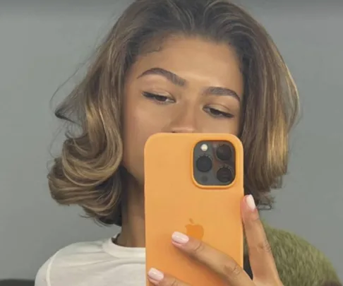 Woman with a blonde bob hairstyle takes a mirror selfie with an orange phone case.