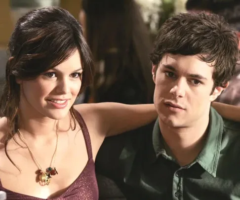 A man and woman sitting closely with her arm around him, both looking in the same direction, in a casual setting.
