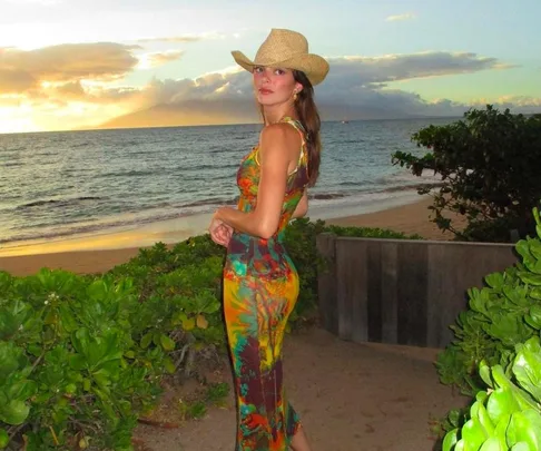 Woman in a cowboy hat and floral dress standing on a beach at sunset.
