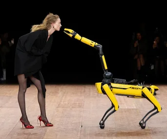 Model leans towards a yellow robotic dog on a wooden runway, wearing a black dress and red heels.