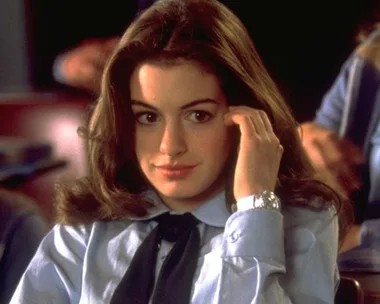 Young woman in a blue shirt and tie sitting in a classroom, with long brown hair, looks thoughtful.