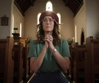 A woman in glasses and a red beret prays in a church with hands clasped, light streaming from windows behind her.