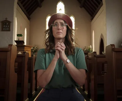 A woman in glasses and a red beret prays in a church with hands clasped, light streaming from windows behind her.