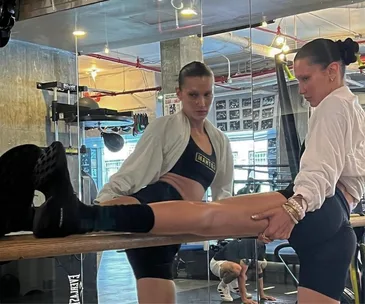 Two women stretching by a mirror in a gym, with workout equipment in the background.