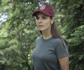 A woman in a maroon cap and gray shirt is standing in a lush forest, looking intently ahead.