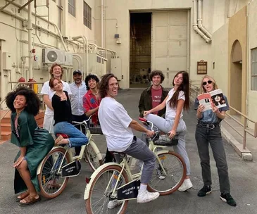 Cast of "Daisy Jones & The Six" smiling, posing with bikes outside a film studio.