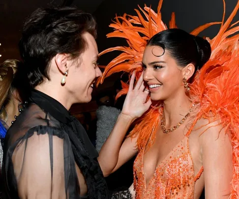 Two people smiling and interacting at an event; one wears a sheer top, the other an orange feathered gown.