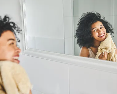 Woman smiling in bathroom mirror, drying face with towel, curly hair.