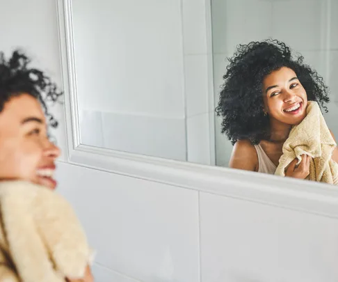 Woman smiling in bathroom mirror, drying face with towel, curly hair.