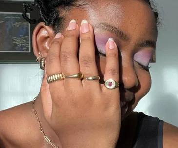 A woman smiling with purple eyeshadow, wearing multiple stacked gold rings on her hand covering her face.