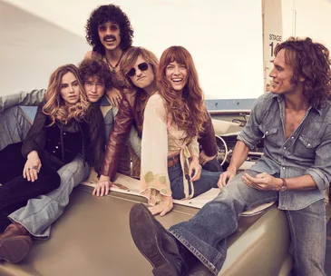 Group of six people in 70s fashion sitting on a car, smiling and relaxed, with a backdrop of a stage area.