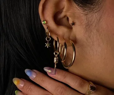 Close-up of an ear with gold helix piercings and star jewelry; hand with colorful nails touches hair.