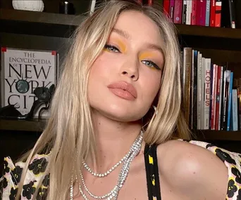 Gigi Hadid poses in front of a bookshelf with colorful eyeshadow and layered necklaces.