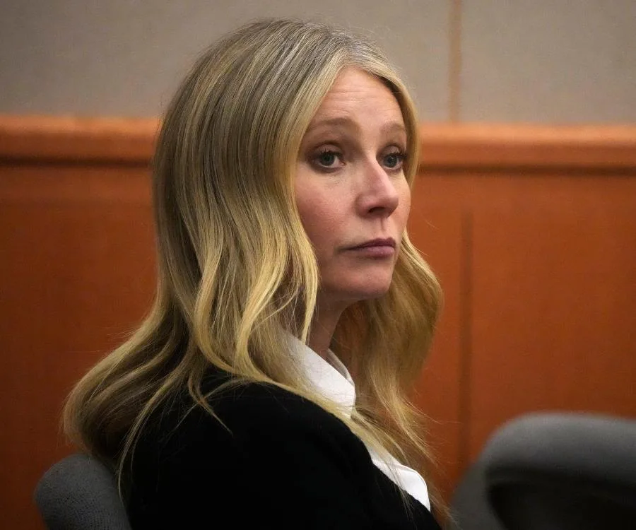 A woman with long blonde hair sits in a courtroom, wearing a black outfit with a white blouse.