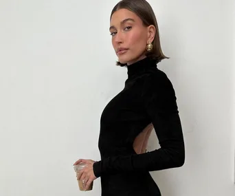 Woman with bob haircut in black backless dress holding drink, poses against white background.