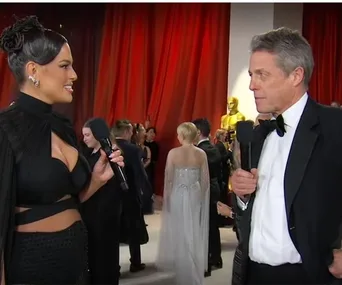 TV host interviewing a man in a tuxedo on a red carpet during an awards event, with people and a gold statue in the background.