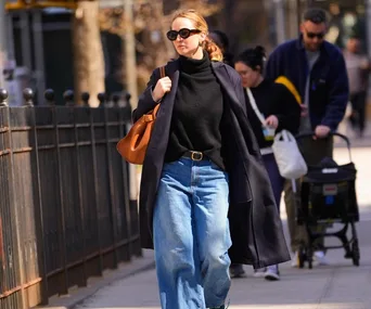 A woman in sunglasses, black coat, and wide-leg jeans walks confidently on a city sidewalk carrying a brown bag.