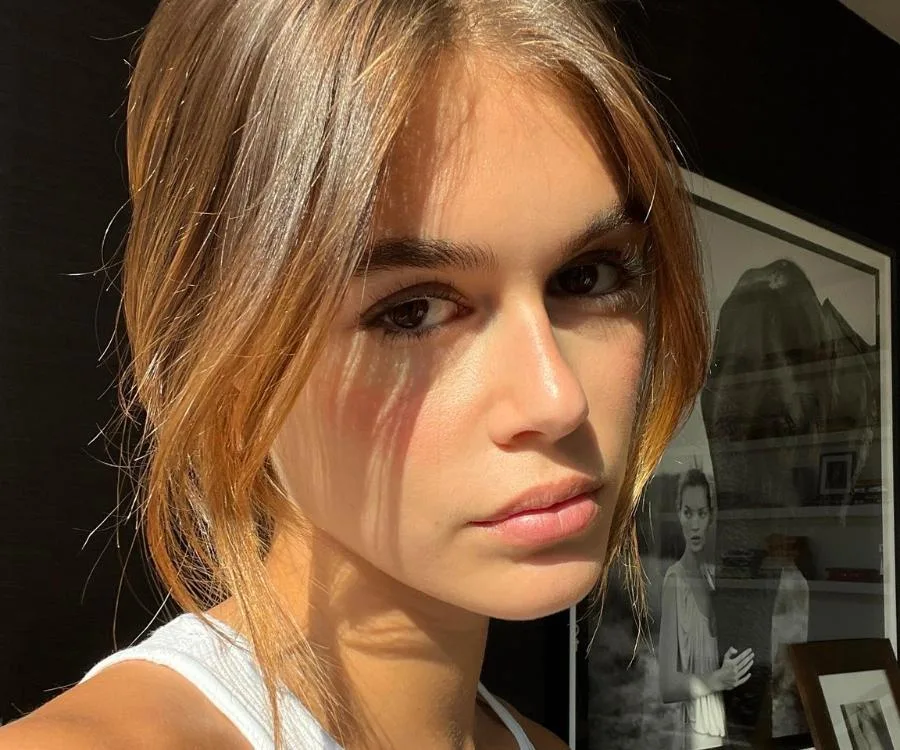 Woman with light brown hair and white top poses by a framed photo; sunlight casts shadows on her face.