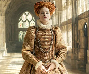 A person dressed as Queen Elizabeth I in an ornate gown with red hair, standing in a historic hallway with stained glass windows.