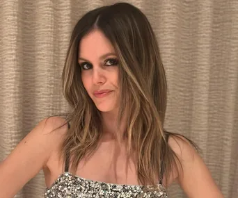 A woman with long brown hair wearing a sparkly top poses indoors with a neutral expression.