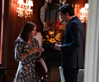 Woman in floral dress holding a Burberry bag talks to man in a suit; elegant room with flowers and lamps in background.