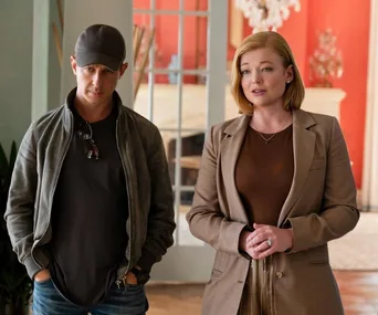 Two characters from Succession, Kendall in a cap and jacket, Shiv in a brown suit, stand in a room with red walls.