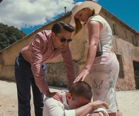 Two people in summer attire console a person crouching on the ground outside a rustic building.