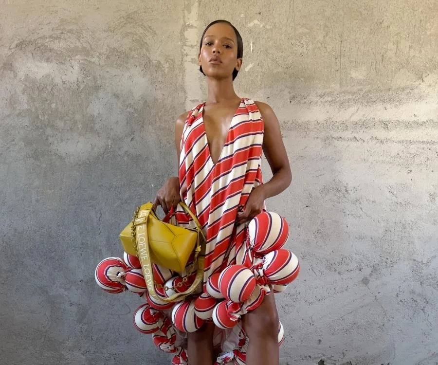 Person in a striped, ruffled dress holds a yellow Loewe bag in front of a textured wall.