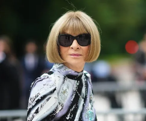 A person with a bob haircut and sunglasses wearing a patterned outfit, outdoors with a blurred background.