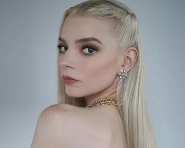 Portrait of a woman with long, straight, blonde hair, looking over her bare shoulder, wearing a floral earring and gold necklace.