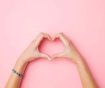 Hands forming a heart shape against a pink background, one wearing a blue flower bracelet.