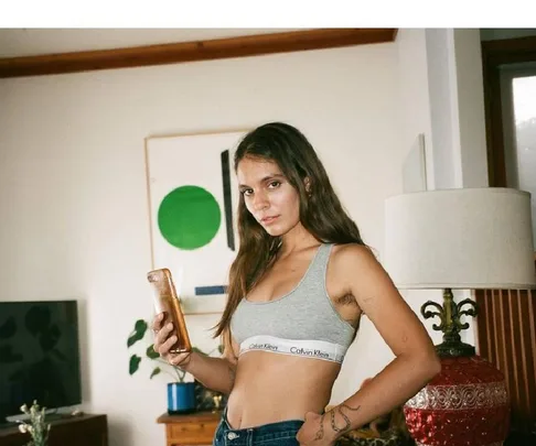 A woman in a gray Calvin Klein sports bra, holding a phone in a cozy, lamp-lit room with art on the wall.