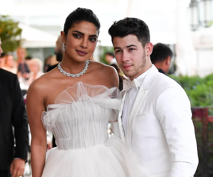 A couple dressed elegantly at an event; the woman in a white gown, jeweled necklace, the man in a white tuxedo.