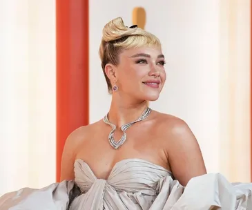 Florence Pugh at the Oscars in a strapless gown with unique hairstyle and diamond necklace, smiling confidently.