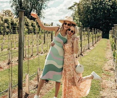 Two women in summer dresses and sunglasses pose happily in a vineyard, one holding a wine glass.