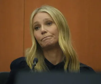 Person with long blonde hair at a microphone in court, looking contemplative.