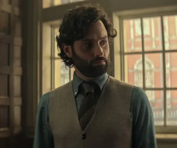 Man with dark curly hair and beard in a vest and tie, standing by a window with a serious expression.