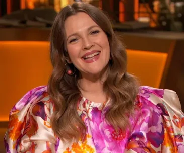 Smiling woman with long hair in a vibrant, floral blouse against a warm, orange-lit background.