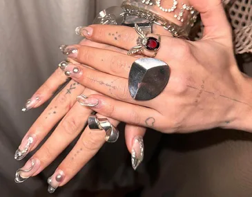 Close-up of hands with silver rings and 3D metallic nail art featuring pearls and abstract designs.