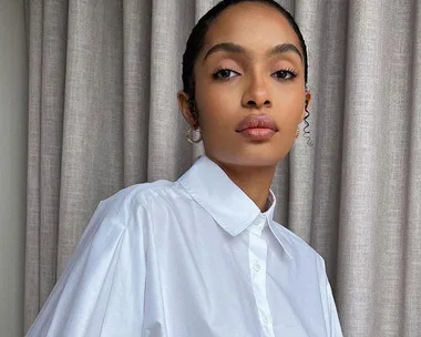 A woman in a white shirt poses confidently in front of beige curtains.