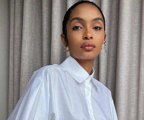 A woman in a white shirt poses confidently in front of beige curtains.