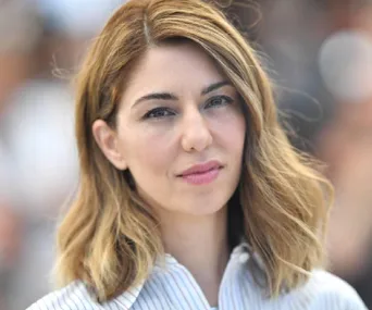 Woman with shoulder-length hair, wearing a striped shirt, looking at the camera during an outdoor event.