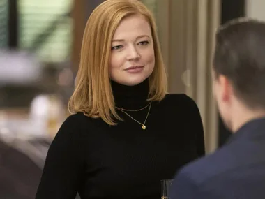 A woman with red hair in a black turtleneck, wearing a necklace, holds a glass while talking to a man in a suit.