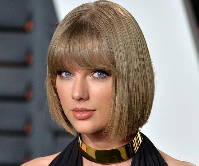 A person with a bob haircut and bangs, wearing a gold choker and black outfit, posing at an event.