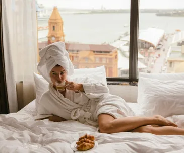 Woman in a bathrobe enjoys coffee on a bed with a city view, croissants nearby on a plate.