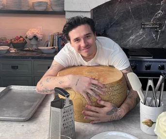 Person hugging a large cheese wheel in a kitchen, with a cheese grater and utensils on the counter.