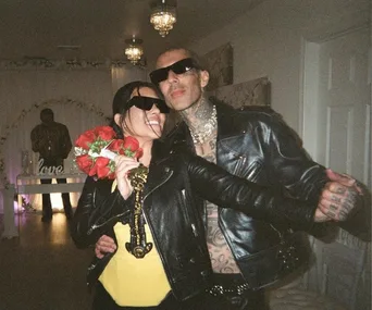 A couple in black leather jackets, sunglasses, and tattoos, holding a bouquet, smiling in an indoor setting.