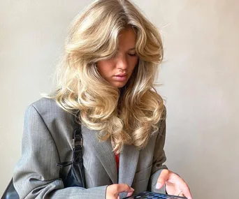 Woman with long blonde hair in a gray blazer looks down at her phone.