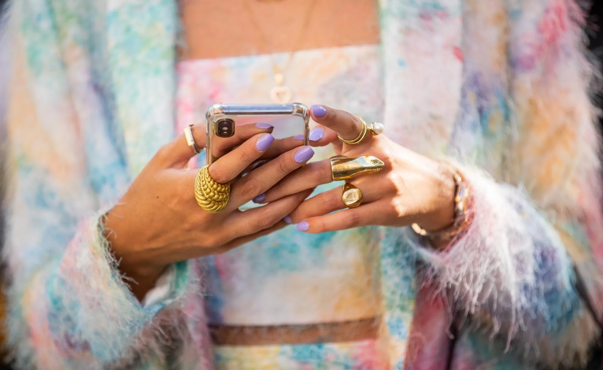 Person with pastel nail polish and chunky gold rings using a smartphone, wearing a colorful, fluffy jacket.